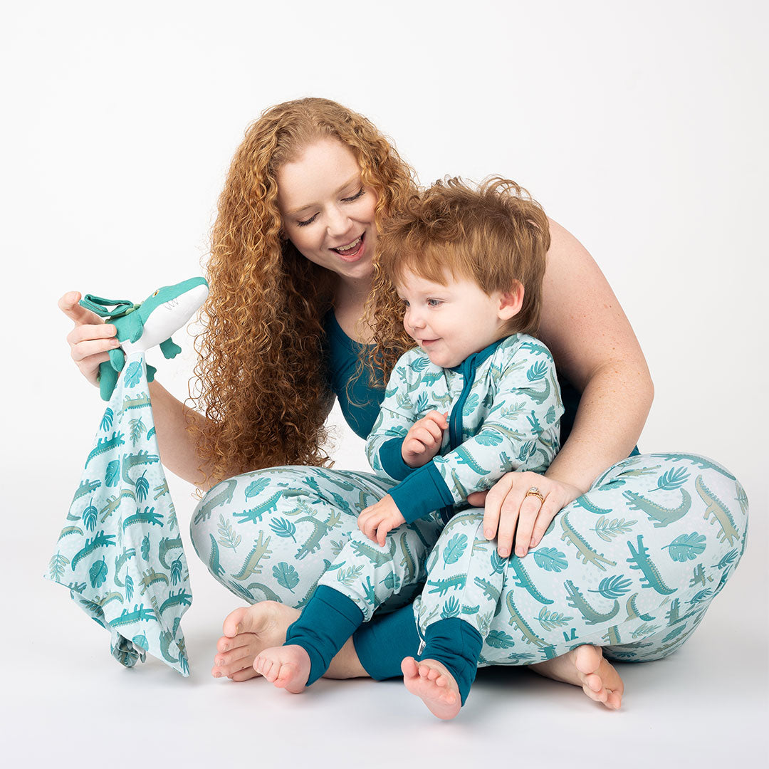 A mother with her son in her lap. they are both sporting different attire in the "later gator" prints. they are playing with the "later gator" lovey. the "later gator" print has a mix of light and dark green alligators, leaves, and white dots scattered on a teal colored background. 
