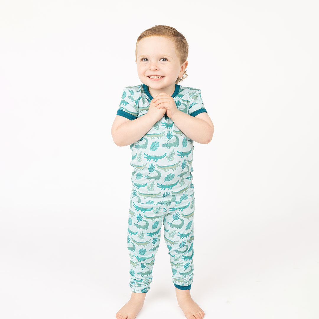 boy smiling in his "later gator" 2-piece matching pajama set. the "later gator" print has a mix of light and dark green alligators, leaves, and white dots scattered on a teal colored background. 