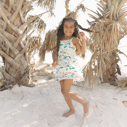Toddler girl smiling while having fun at the beach in Emerson and Friends Coral Reef Bamboo Sundress, featuring a soft, breathable fabric with an ocean-themed print of sharks, sea turtles, and coral, designed for lightweight summer comfort and easy movement.
