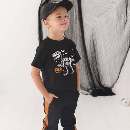 A young boy wearing a black t-shirt with a white skeleton dinosaur and orange pumpkin graphic, paired with black pants featuring orange side stripes, a black cap, and standing in front of a black spiderweb backdrop.