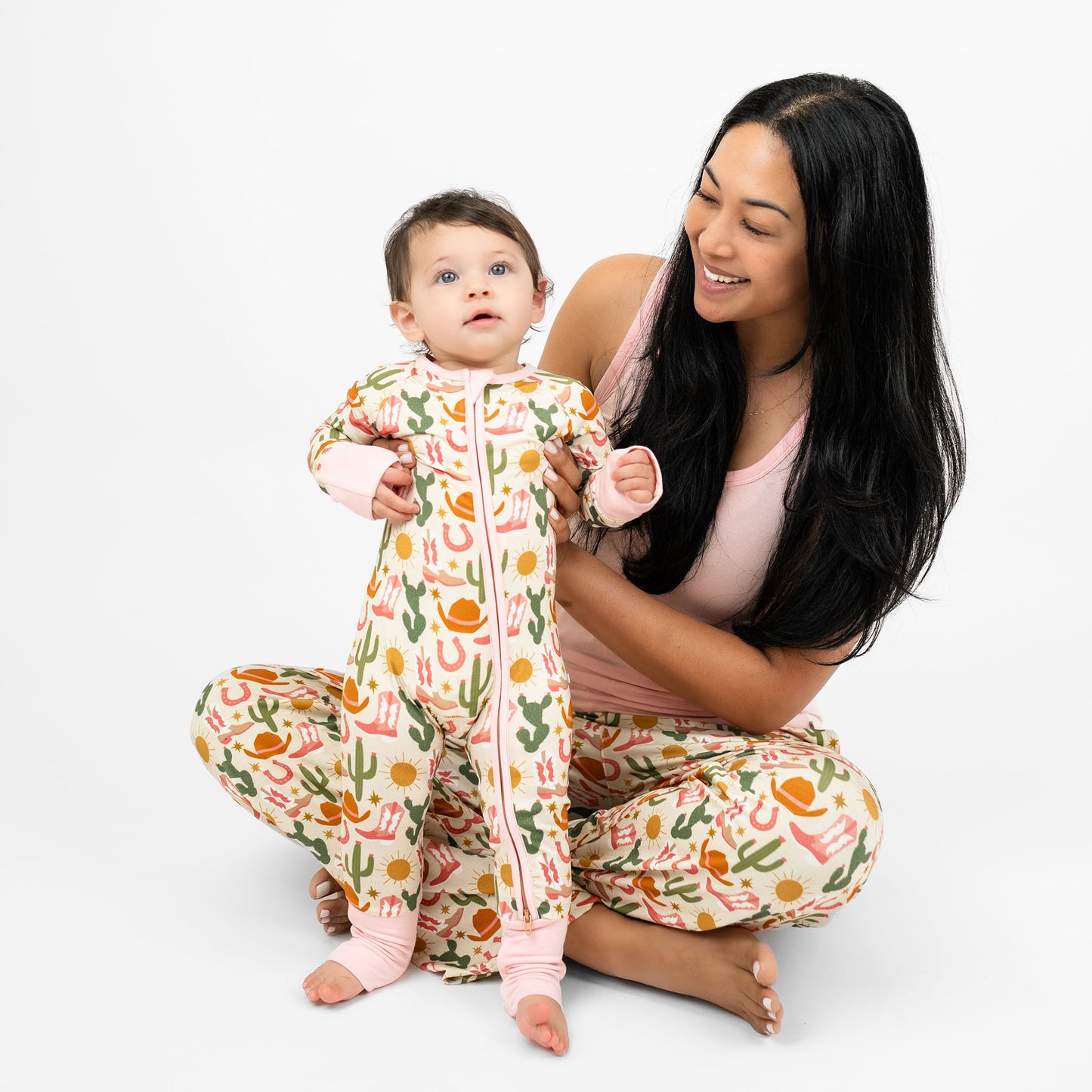 Baby girl and mom wearing our Emerson and Friends Hey Y’all Convertible Bamboo Convertible Pajamas and Hey Y'all relaxed pants. The print shows  green cactus, yellow sun, brown cowgirl hat, pink boots and horseshoes. Perfect for cozy rodeo nights or pajama parties with a cowgirl flair!