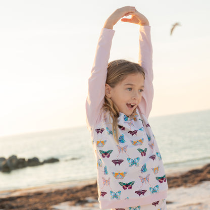 Young girl with arms raised on a beach wearing a butterfly-patterned rashguard.